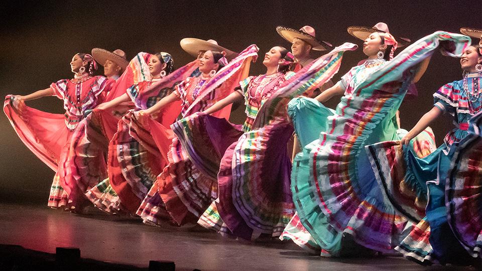 six dancers in colorful, traditional dresses lift their skirts in a semi-circle